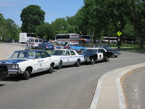 1991 Ford LTD Crown Victoria - Several vintage New York State Police and other marked police cars are lined up along a tree-lined road with buses behind.