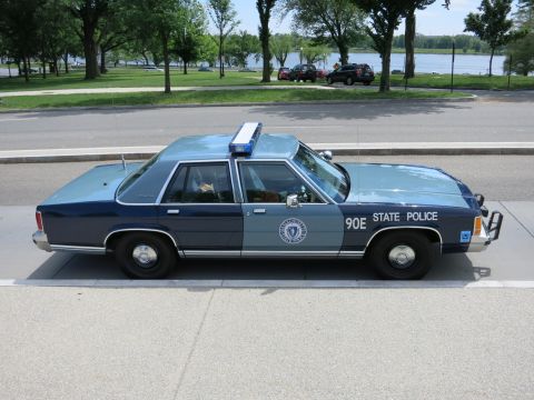 1991 Ford LTD Crown Victoria - two-tone blue '90E State Police' sedan sits in profile along a curb with a river and parked cars behind.