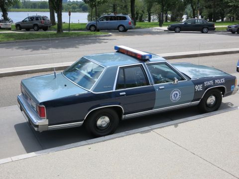 1991 Ford LTD Crown Victoria - Rear three-quarter view of a blue '90E State Police' sedan with red-and-blue light bar parked near a riverside park.