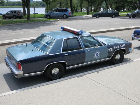 1991 Ford LTD Crown Victoria - blue '90E State Police' sedan is parked rear three-quarter at a curb along a waterfront roadway.