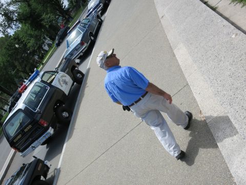 1991 Ford LTD Crown Victoria - man in a blue shirt and white cap walks along a sidewalk past a line of parked black-and-white police cars.
