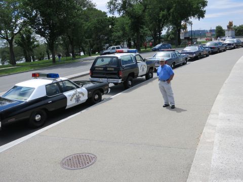 1991 Ford LTD Crown Victoria - man in a blue shirt stands on a wide sidewalk beside a long row of parked vintage police cars and an SUV.