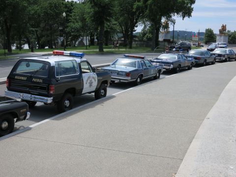 1991 Ford LTD Crown Victoria - black-and-white Highway Patrol SUV leads a line of parked vintage police sedans along a curving roadway.