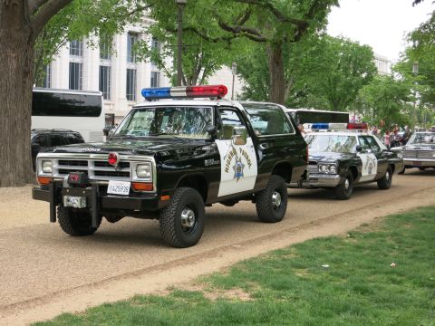 1991 Ford LTD Crown Victoria - black-and-white Highway Patrol Dodge Ramcharger SUV with light bar and gold star emblem, front three-quarter, parked on grass by a building.
