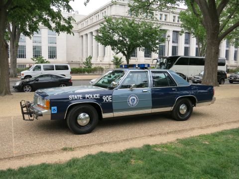 1991 Ford LTD Crown Victoria - two-tone blue State Police Ford LTD Crown Victoria sedan with push bar and seal, marked STATE POLICE 90E, parked under trees by a stone bui…