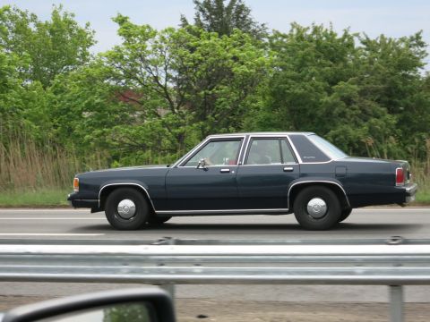 1991 Ford LTD Crown Victoria - dark blue Ford LTD Crown Victoria sedan moving along a highway past green roadside foliage, shot from another vehicle.