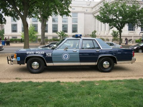 1991 Ford LTD Crown Victoria - Side profile of a two-tone blue State Police Ford Crown Victoria marked STATE POLICE 90E with a push bar, parked before a large stone build…