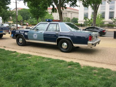 1991 Ford LTD Crown Victoria - Rear three-quarter view of a two-tone blue State Police Ford Crown Victoria marked STATE POLICE 90E, parked on a dirt verge by a city build…
