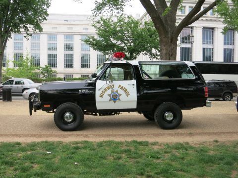 1991 Ford LTD Crown Victoria - Side profile of a black-and-white Highway Patrol Dodge Ramcharger SUV with light bar and gold star emblem, parked on a dirt path by trees.