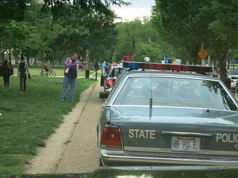 1991 Ford LTD Crown Victoria - Rear of a dark green State Police Ford sedan marked STATE POLICE with a Massachusetts plate 90E, as people stand on a park path nearby.