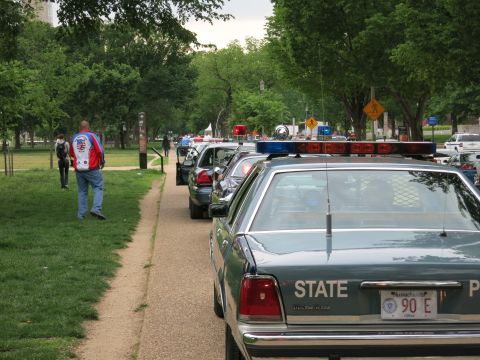 1991 Ford LTD Crown Victoria - row of police cars parked along a tree-lined park path, led by a dark State Police Ford sedan with light bar and Massachusetts plate 90E.