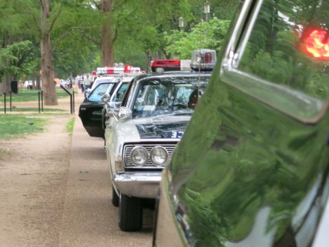 1991 Ford LTD Crown Victoria - line of vintage police cars parked along a dirt park path, led by a chrome dual-headlight sedan, with red emergency lights lit behind.
