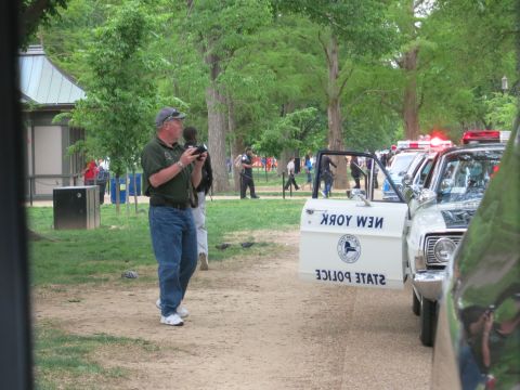 1991 Ford LTD Crown Victoria - man in a baseball cap and green polo holds a camera near a row of police cars, including a New York State Police vehicle, parked along a tr…