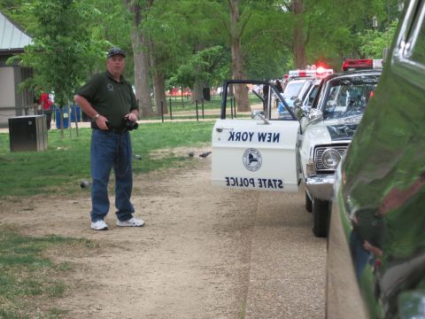 1991 Ford LTD Crown Victoria - bearded man in a green polo and cap stands holding a camera beside a white New York State Police cruiser with flashing lights, on a park pa…