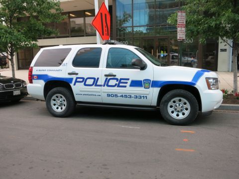 1991 Ford LTD Crown Victoria - white and blue Peel Police Chevrolet Tahoe SUV sits curbside near a dark BMW, in front of a glass-fronted downtown building.