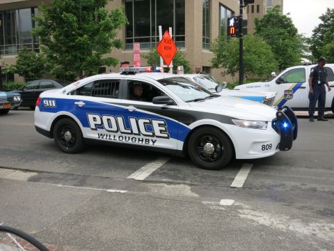 1991 Ford LTD Crown Victoria - white and blue Willoughby Police Ford Taurus Interceptor with flashing lights navigates a city street as an officer stands nearby.