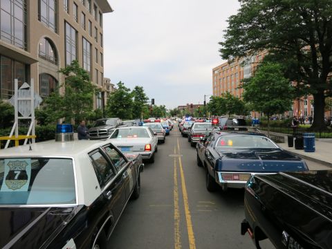 1991 Ford LTD Crown Victoria - Rows of classic police cars, including black-and-white cruisers, fill a downtown street between office buildings in a parade-like processio…