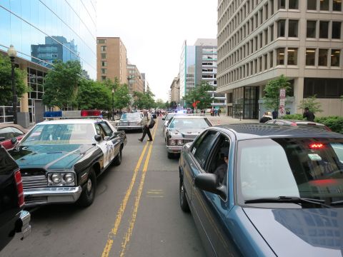 1991 Ford LTD Crown Victoria - black-and-white vintage police cruiser leads a line of patrol cars down a wide city avenue lined with modern office buildings.