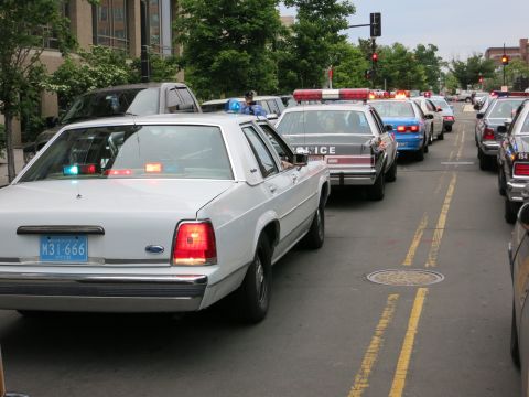 1991 Ford LTD Crown Victoria - white Ford LTD Crown Victoria police car with lit brake lights and a New York plate waits in a line of patrol cars on a city street.