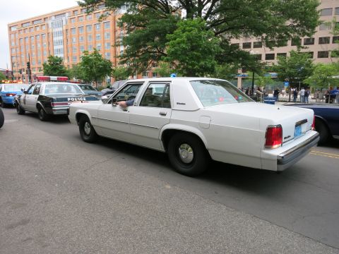 1991 Ford LTD Crown Victoria - white Ford LTD Crown Victoria police sedan drives down a city street with other marked cruisers and pedestrians in the background.