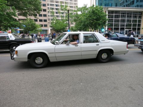 1991 Ford LTD Crown Victoria - white Ford LTD Crown Victoria police sedan with a driver and passenger cruises a downtown street past a crowd and other patrol cars.