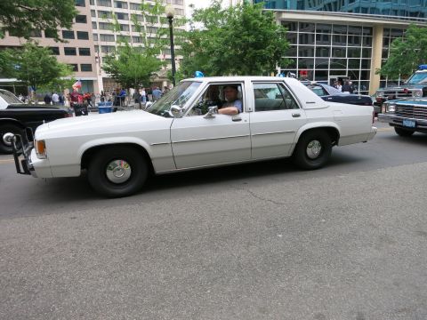 1991 Ford LTD Crown Victoria - white Ford LTD Crown Victoria police car with occupants drives along a downtown street lined with spectators and other cruisers.