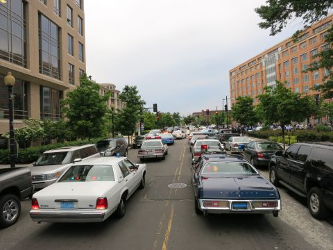 1991 Ford LTD Crown Victoria - elevated view shows white and vintage police cars, including a wagon, filling both lanes of a wide downtown avenue between brick buildings.