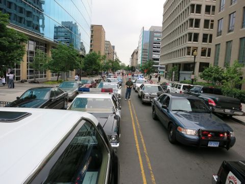 1991 Ford LTD Crown Victoria - man stands on the centerline of a downtown street flanked by rows of parked police cruisers under glass and concrete office towers.