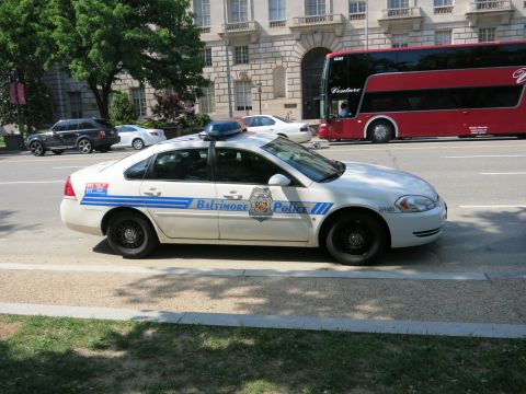 1991 Ford LTD Crown Victoria - white Baltimore Police Chevrolet Impala cruiser is parked curbside near a stone government building as a red double-decker bus passes.
