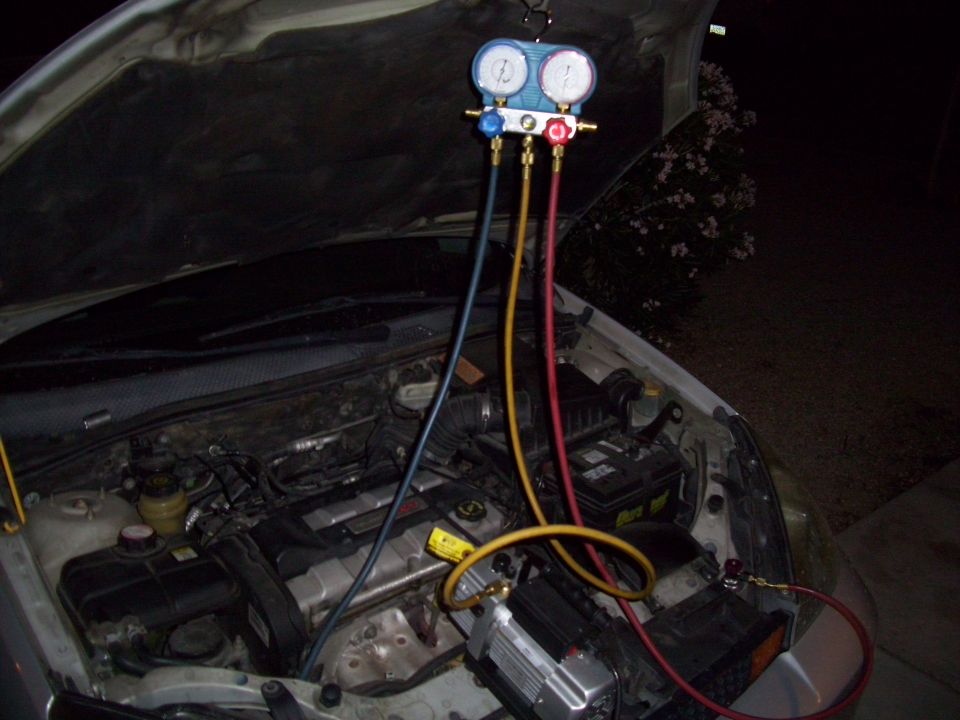 Car engine bay with AC manifold gauges connected to the system. A vacuum pump is also attached
