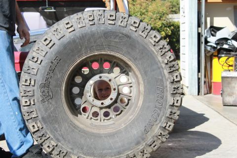 1968 Ford Bronco - A child's face is visible through the spokes of a large tire, with a red vehicle partially seen behind it.