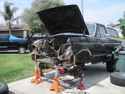 1989 Ford Bronco - The front end of a black Ford Bronco is dismantled and elevated on jack stands, revealing its chassis and suspension components.