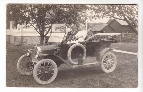 1909 Ford Model T - A couple poses in a 1909 Ford Model T convertible on a grassy lawn with a house and barn in the background.