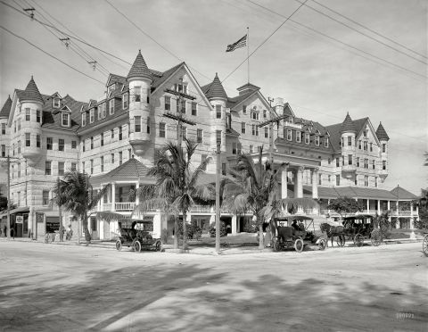 1909 Ford Model T - A 1909 Ford Model T is seen parked on a street in front of a grand hotel, with a horse-drawn carriage also visible.