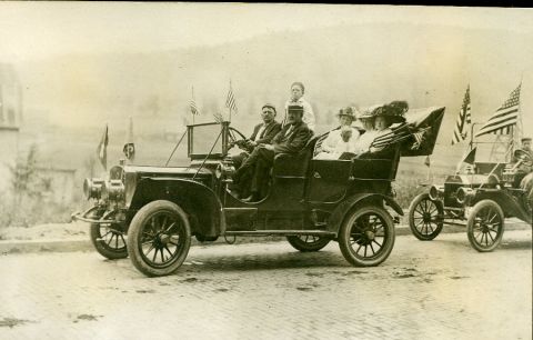 1909 Ford Model T - Passengers ride in a touring car, with another vehicle visible behind it, during what appears to be a parade or celebration.