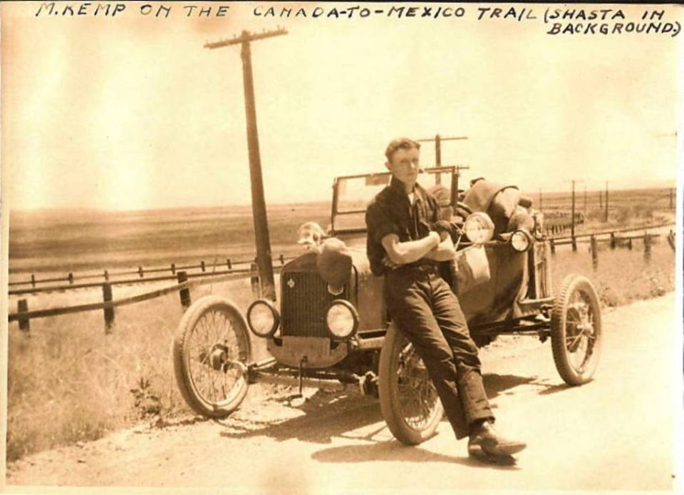 Man leaning on a dark-colored 1910 automobile with a wooden fence and telephone pole nearby