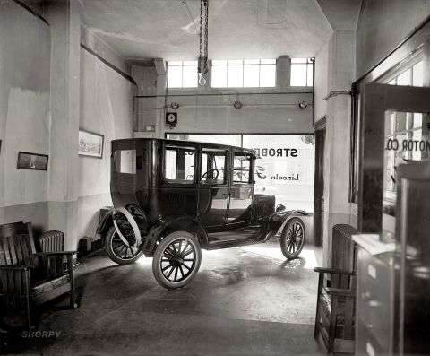1909 Ford Model T - A dark sedan is parked inside a garage, with windows showing a reflection of a sign that reads "MOTOR CO" and "Lincoln".