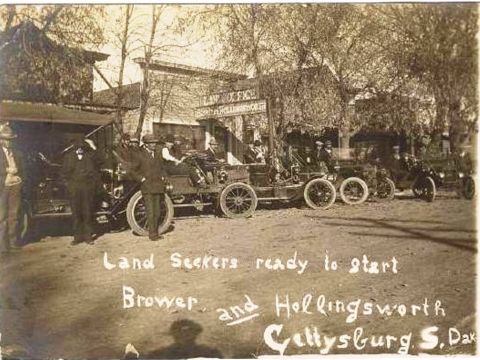 1909 Ford Model T - Multiple early automobiles are lined up on a dirt road, with people gathered around them. Text indicates a 'Land Seekers' event.