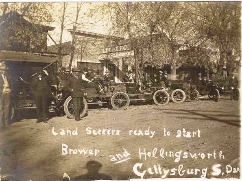 Several early automobiles parked on a dirt road in front of buildings, with people standing around them. Text reads 'Land Seekers ready to start Brower and Hollingsworth Celly'