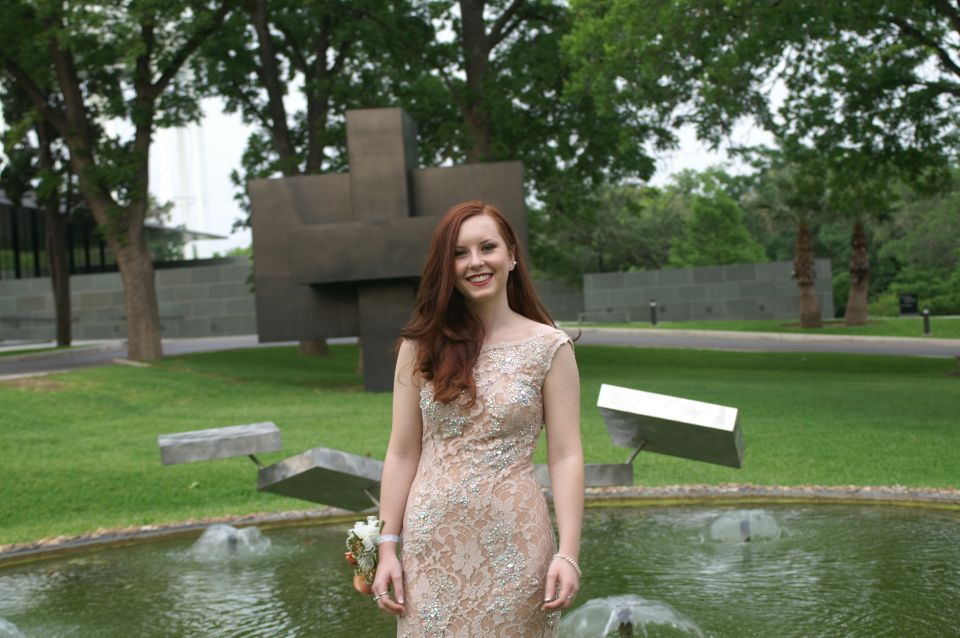 Woman in a light-colored, beaded formal dress stands in front of a fountain and modern sculpture