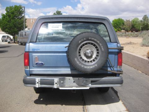 1988 Ford Bronco - This blue Ford Bronco is shown from the rear, a spare tire mounted externally. A sticker is visible on the back window.