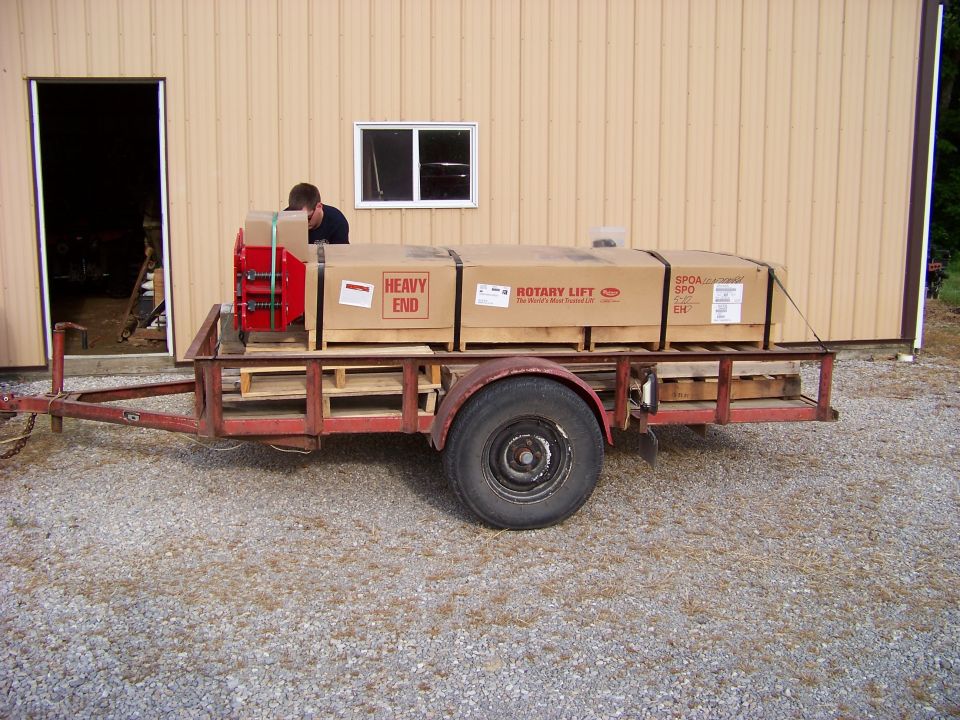 Red trailer loaded with cardboard boxes and a red piece of equipment, parked on gravel in front of a tan building