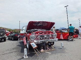2013 Ford F-100 - A red pickup truck is displayed with its hood open, patriotic graphics, at an outdoor car show with other vehicles present.