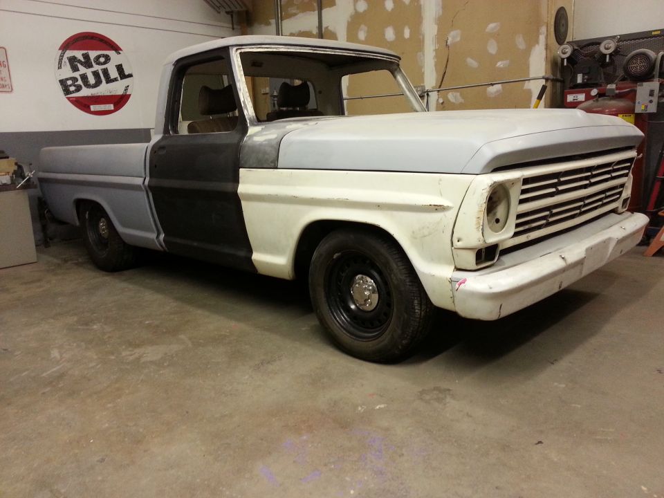 Front 3/4 view of a 1967 Ford F-100 pickup truck in a garage, primer gray and black paint, undergoing restoration