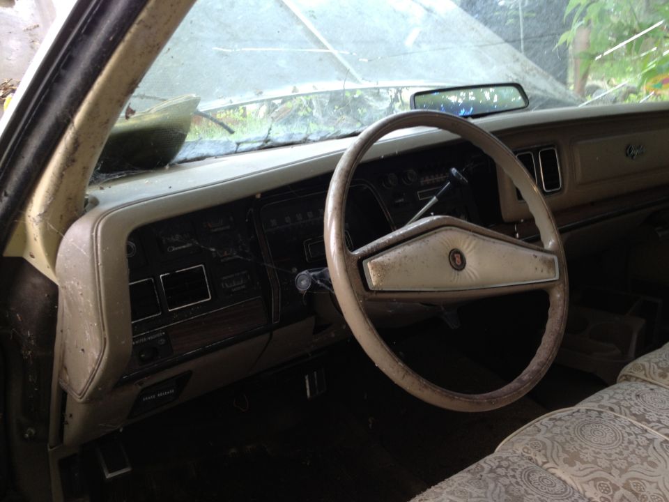 Interior view of an old car's dashboard and steering wheel. The dashboard has wood grain accents and rectangular vents