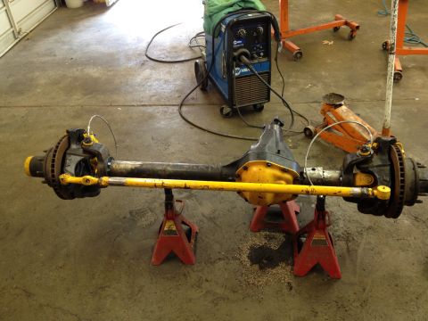 1988 Ford Bronco - Front axle assembly with yellow tie rod, supported by red jack stands, with a welding machine in the background.