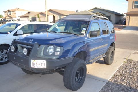 2004 Nissan Xterra - A blue Nissan Xterra is parked in a residential driveway, showing its front-left side and a portion of a white SUV.