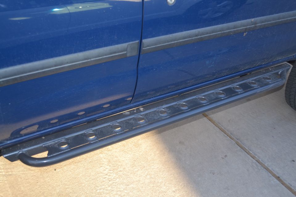 Side of a blue SUV showing the door and aftermarket running boards with circular cutouts. A tire is visible on the right