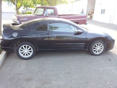 2001 Mitsubishi Eclipse - A black coupe with a spoiler and peeling paint is parked next to a maroon pickup truck.
