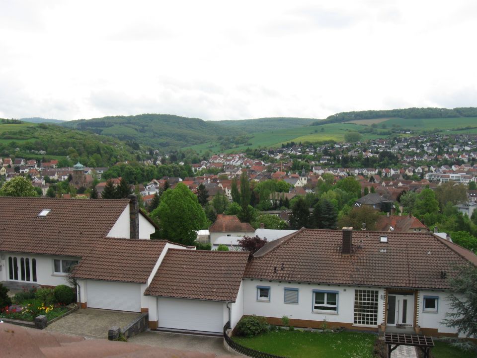 View of a suburban town with houses and rolling green hills under a cloudy sky. Watermark reads SUPERMOTORS.NET